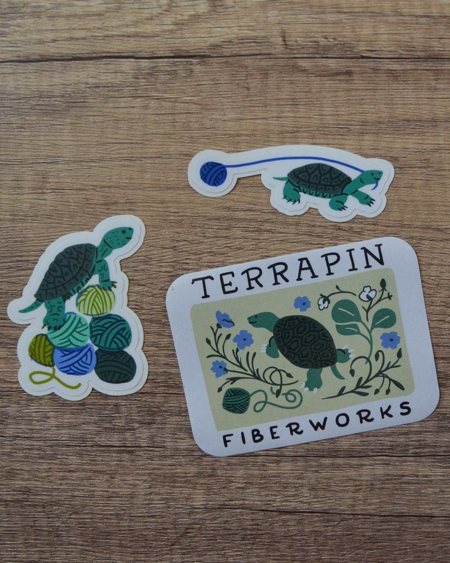 Three stickers featuring turtles and yarn on a wooden surface, with 'Terrapin Fiberworks' branding.